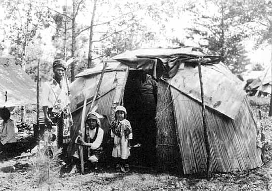 Chippewa family standing in front of birch bark and reed wigwam,