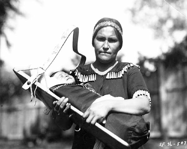 Ojibway woman wearing jingle dress holding baby in cradle board, 1936