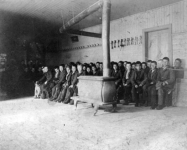 Young boys seated by wood stove, Indian boarding school, location unknown, ca. 1900.