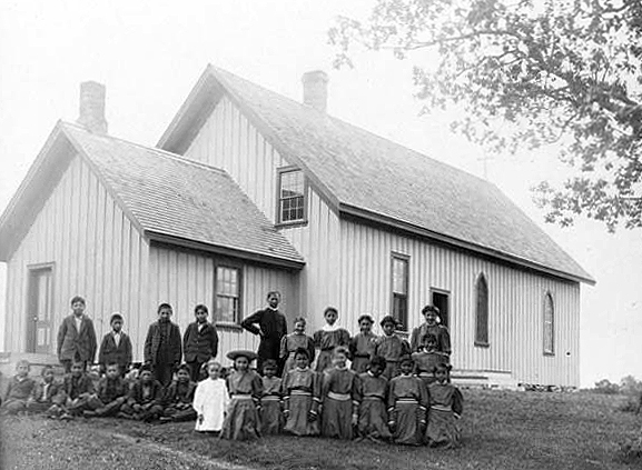 Priest posed with children in front of church, Indian boarding school, location unknown, ca. 1900.